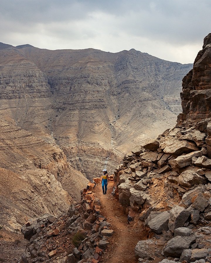 woman on a hiking trail in uae mountains wearing blue leggings and white cap on a cloudy evening