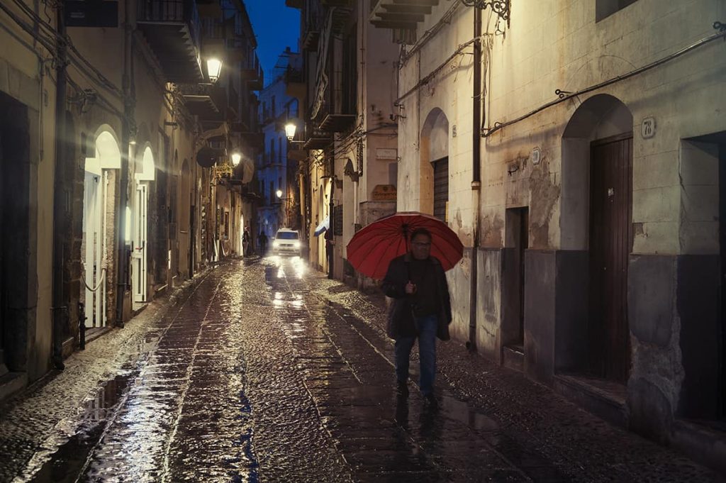 a man with red umbrella walking on a rainy street in cefalu at night, sicily