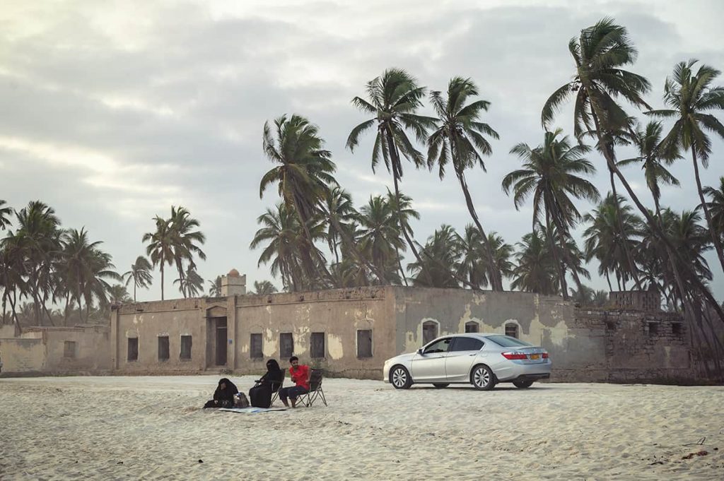 Salalah city beach with palmtrees and old mosque with locals sitting on the sand