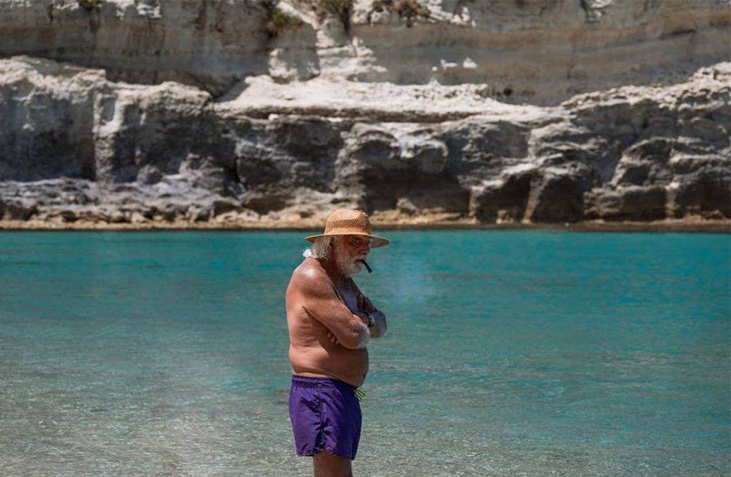 a man with cigar and a hat waring purple swimming shorts in tropea by the beach 