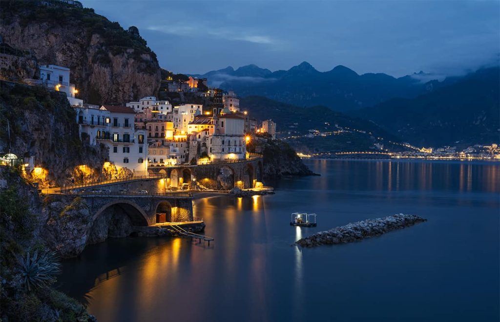 Night descends on Amalfi, a gem among the best towns on the Amalfi Coast, with warm lights illuminating the historic buildings and arches along the coastline. The serene sea reflects the lights, and a solitary boat glides by, against a backdrop of distant mountains under a dusky sky.