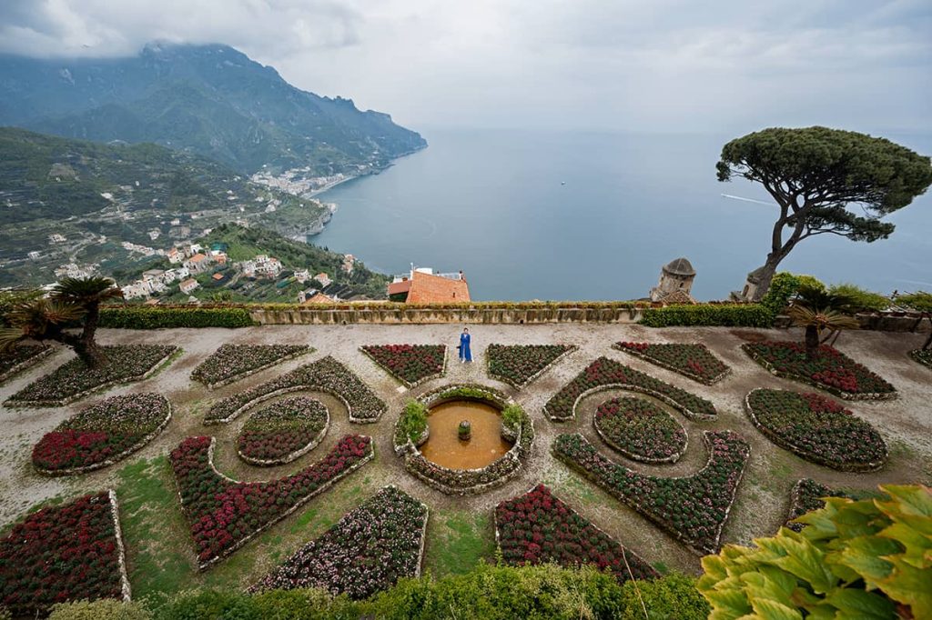 Elevated view of the formal gardens at Villa Rufolo in Ravello, one of the best towns on the Amalfi Coast, with geometric hedge patterns and colorful flower beds overlooking the panoramic vista of the coastline and the Tyrrhenian Sea, under a dramatic, cloud-filled sky. A woman in blue dress is standing on the terrace.