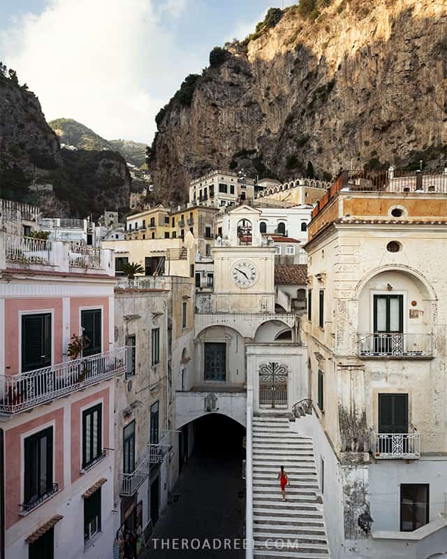 A tranquil street in Atrani, featuring the historic architecture of white and pastel-colored buildings with a staircase leading to an arched tunnel, set against the dramatic backdrop of steep cliffs and lush greenery.