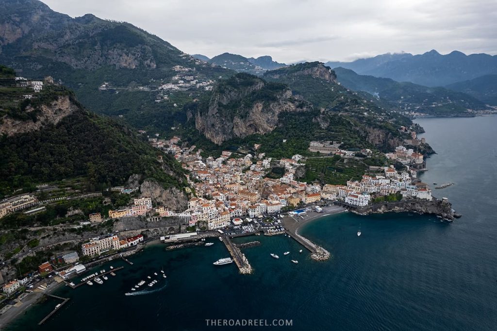 Sweeping aerial view of the coastal town of Amalfi, snuggled between the cliffs and the sea, with its dense cluster of white buildings, the marina filled with boats, and the open arms of the Mediterranean, all under the watchful presence of the lush Lattari Mountains.