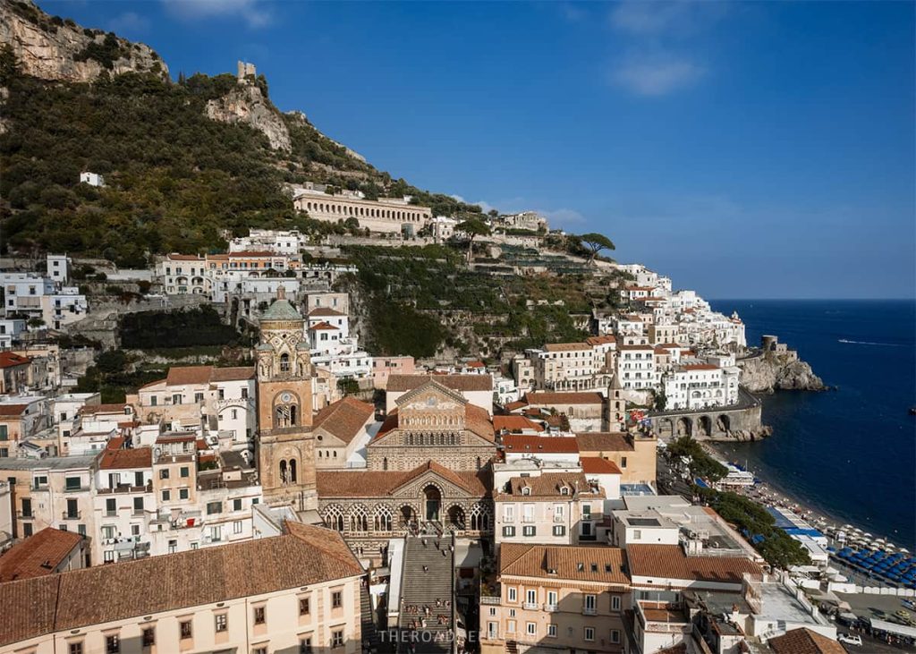 Panoramic view of Amalfi town from above, with the grand Cathedral of Saint Andrew and its ornate facade in the center, surrounded by a dense layout of white buildings against the cliffside, leading to the clear blue sea.