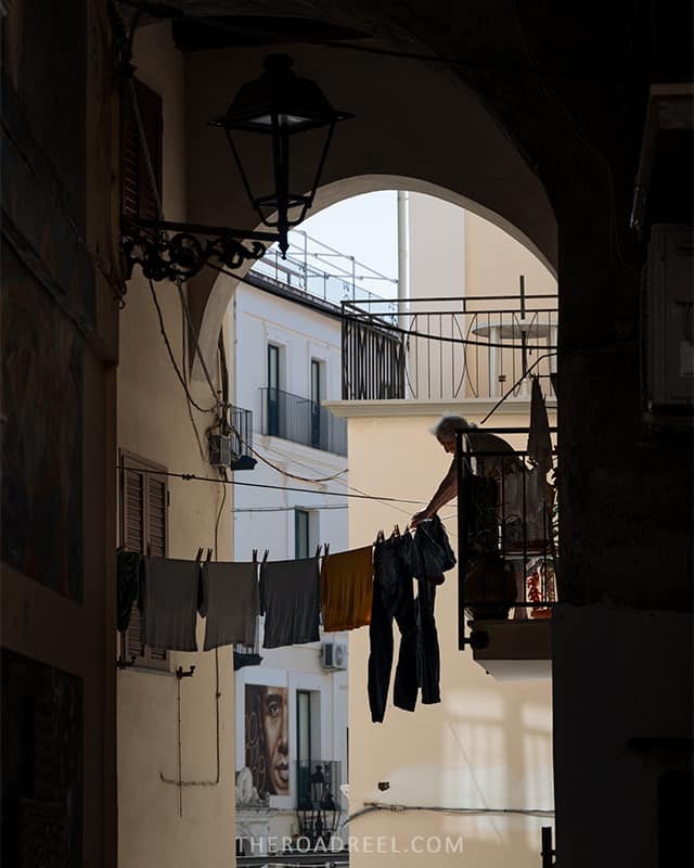 an elderly lady hanging laundry on a rope while standing in a balcony, you can see a mural of Diamante, Calabria in the background