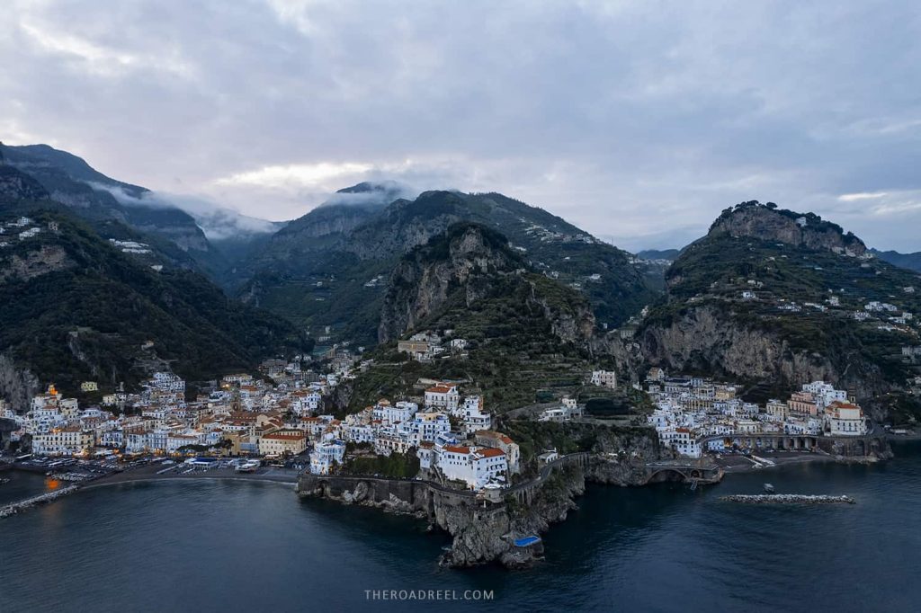 aerial view of Amalfi and Atrani towns at dusk, white washed buildings are dramatically positioned between craggy rocks, and clouds are hivering low touching the mountains