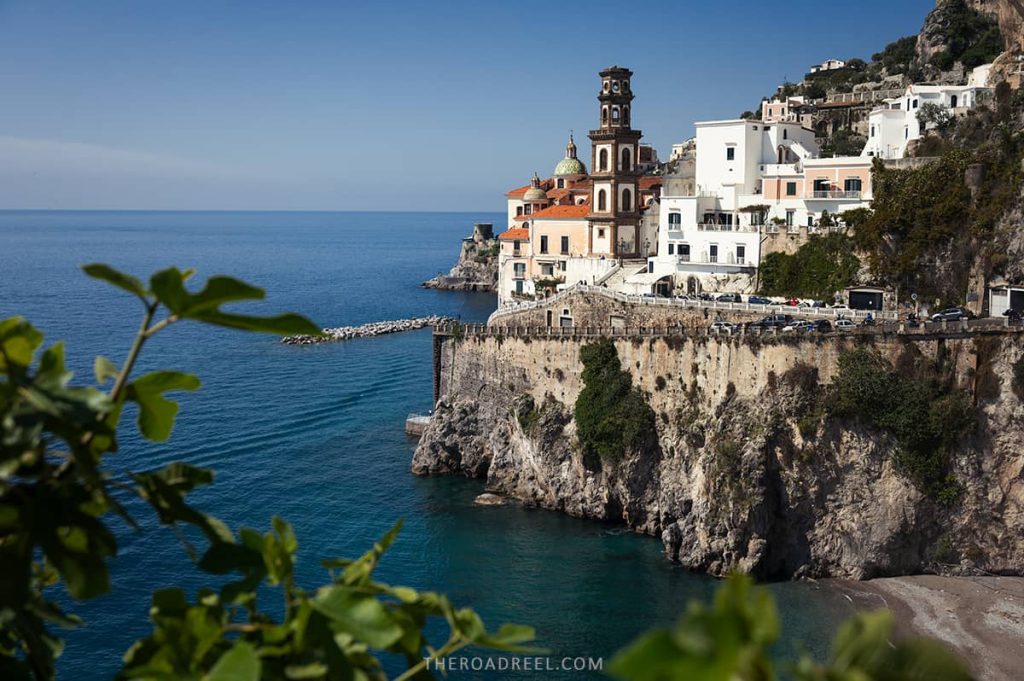 Seaside view of Atrani, the best towns on the Amalfi Coast map, with its signature church bell tower rising above white buildings nestled on the cliff's edge overlooking the serene blue sea, framed by fresh green foliage in the foreground.