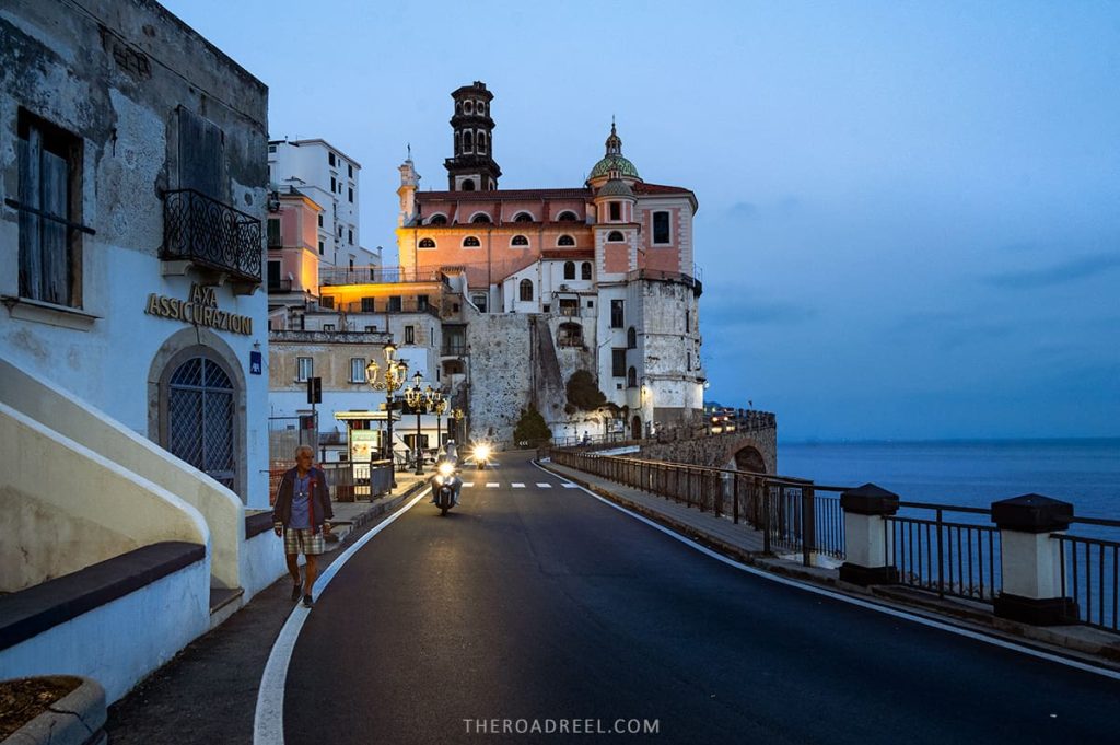 Twilight descends on Atrani, a coastal village on the Amalfi Coast, where the street lamps cast a warm glow on the historic buildings and the impressive architecture of the Church of San Salvatore de Birecto, while locals and travelers alike enjoy an evening stroll by the serene sea.