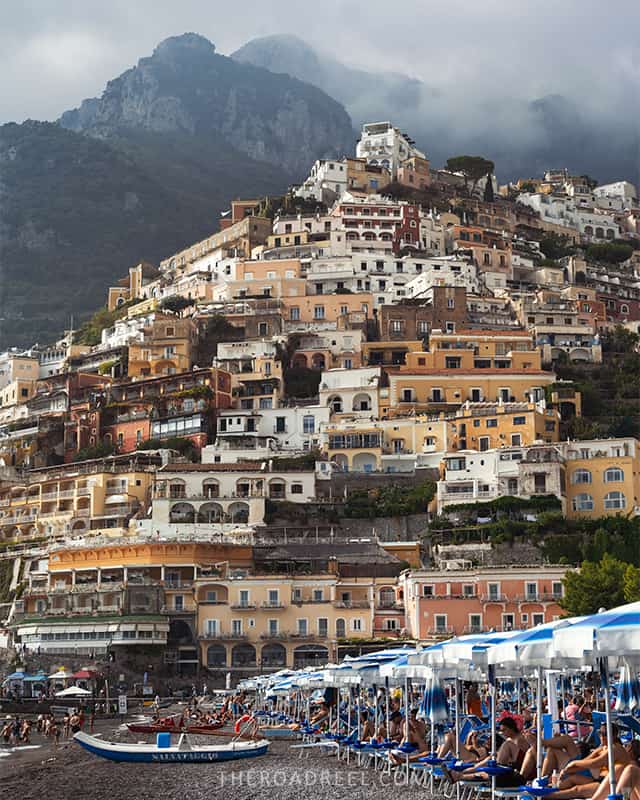 Positano's famous Marina Grande beach buzzes with activity, framed by the town's iconic terraced architecture under a moody sky, capturing why it's celebrated as one of the best towns on the Amalfi Coast