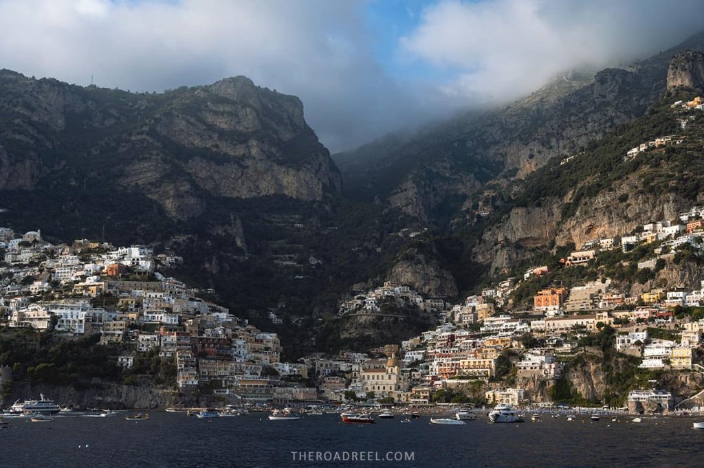 colorful houses of positano, a town on the amalfi coast, dramatically cascading down to the sea, with jagged cliffs rising above and clouds hovering at the peaks, the view is from a ferry