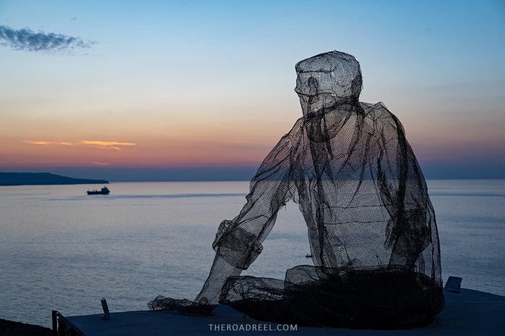 pizzo seaside town in calabria, a huge metal transperent sculpture of a man sitting on the rooftop looking towards the sunset in the sea