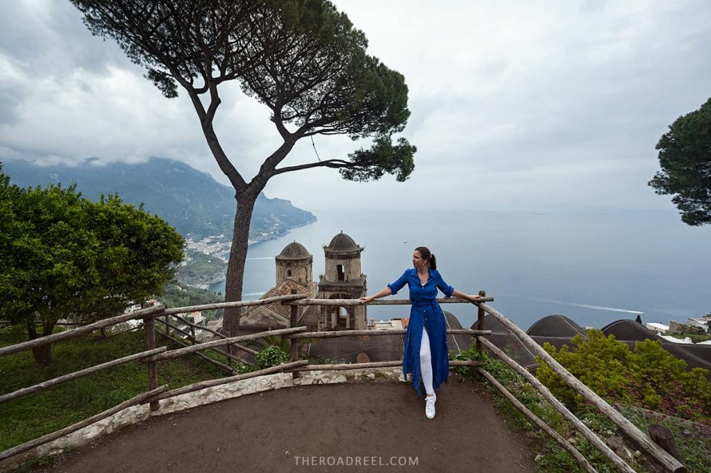 A woman in a blue dress stands at a viewpoint in Ravello, one of the Amalfi Coast's premier towns, with the iconic dome of the Villa Rufolo and the vast expanse of the Tyrrhenian Sea in the background, all framed by a majestic pine tree and the lush coastline.