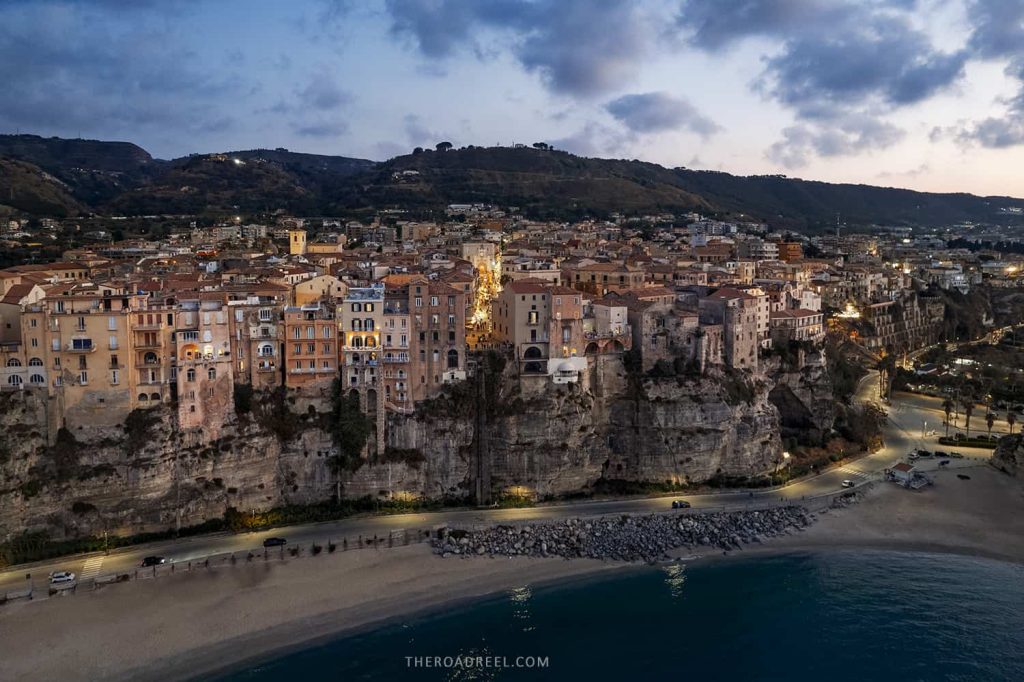 tropea beach town in calabria at dusk from drone, houses clinging above the cliff and tropea beach bellow