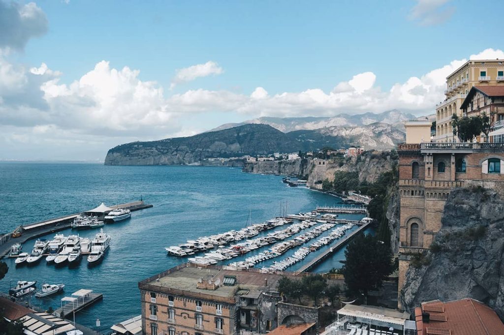 a dramatic coast of Sorrento town, with historic buildings built on the rocks, yackts lining the pier along the sparkling blue sea, beautifuk clouds hovering above the mountains in the background